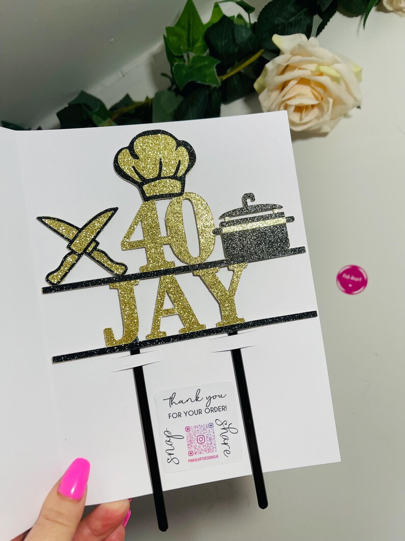 May include: A white card with a gold glitter "40" and "JAY" cake topper. The topper features a chef's hat, crossed knives, and a pot. A small sticker reads "thank you for your order!" with social media details.