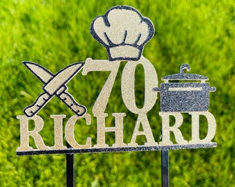 May include: A gold glitter cake topper with the name "RICHARD" and the number "70". The topper includes a chef's hat, crossed knives, and a pot. The background is green grass.