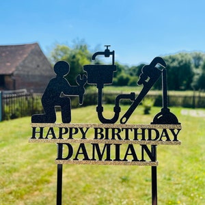 May include: Black and gold cake topper with a silhouette of a plumber holding a wrench and a sink. The text "Happy Birthday Damian" is written on the topper.