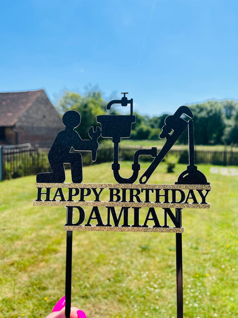 May include: Black and gold cake topper with a silhouette of a plumber holding a wrench and a sink. The text "Happy Birthday Damian" is written on the topper.
