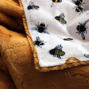 May include: A soft, brown faux fur blanket with a white fleece blanket underneath. The white blanket has a repeating pattern of black and yellow bees.