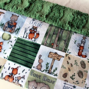 May include: A patchwork baby blanket with a green plush border. The blanket features woodland animal illustrations, including bears, foxes, and rabbits, with the words "Explore", "This Way to Adventure", and "Don't Wake". The blanket is made of soft fabric.