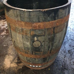 May include: A large wooden barrel with rusty metal bands. The barrel is made of dark wood and has a natural finish. It is sitting on a concrete surface.