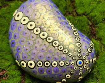 Pode incluir: Uma pedra pintada à mão com um padrão de pontos roxos, brancos e pretos. A pedra é pintada com um desenho de círculos e pontos em espiral. A pedra tem um ponto azul no centro do desenho.