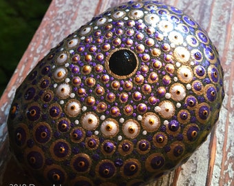 Pode incluir: Uma pedra decorativa pintada à mão com uma base verde escura, adornada com círculos concêntricos de pontos roxos, dourados e brancos. Um grande ponto preto no centro. Uma obra de arte única.