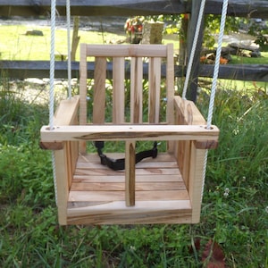 May include: A wooden swing seat with a natural finish and rope hanging straps. The swing is hanging from a tree branch in a grassy area with a wooden fence in the background.