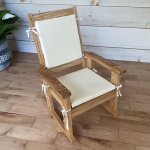 May include: A wooden rocking chair featuring a natural light brown wood grain finish, equipped with two removable off-white fabric cushions. The cushions are secured to the chair frame with matching fabric ties at the corners and sides.