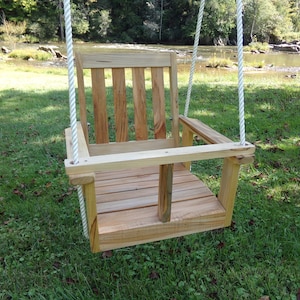 May include: A wooden swing seat with a slatted back and a rope suspension system. The swing is hanging in a grassy area with a river in the background.