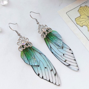 Könnte beinhalten: Ein Paar silberne Ohrringe mit grünen und blauen, schillernden Flügeln. Die Flügel haben ein zartes, mehrschichtiges Design und sind mit Glitzer verziert.