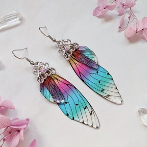 May include: A pair of iridescent wing-shaped earrings. The earrings feature a gradient of colors, including pink, blue, and yellow. The wings are attached to silver-colored metal with decorative filigree and a small, clear gem. The earrings are on a white surface.