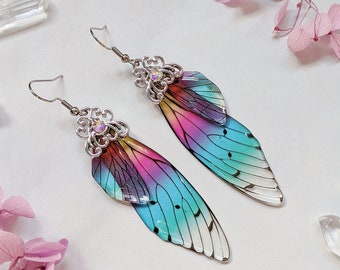 Puede incluir: Un par de pendientes de alas de hadas iridiscentes. Las alas hacen la transición del rosa al azul, con detalles de venas negras. Los pendientes tienen detalles de filigrana plateada y ganchos para las orejas. Cristales y flores secas en el fondo.