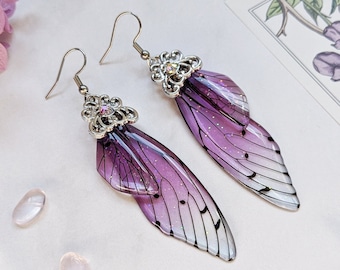 Puede incluir: Un par de pendientes de alas de hada con un degradado de morado y blanco. Las alas están detalladas con venas negras y purpurina. Los pendientes tienen detalles y ganchos de metal plateado.