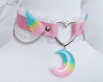 May include: Pink choker necklace with a heart-shaped center and a crescent moon pendant. The choker features rainbow-colored wing accents and a white lace trim. A silver chain hangs from each wing.