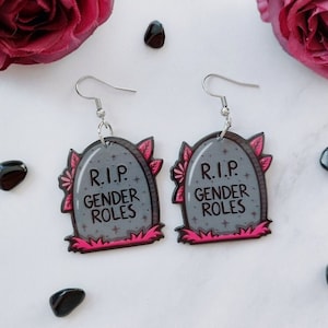 Könnte beinhalten: Ein Paar ausgefallene Ohrringe mit Grabstein-Design. Die grauen Grabsteine tragen den Text "R.I.P. GENDER ROLES" in Weiß, mit rosa und roten floralen Akzenten. Die Ohrringe hängen an silberfarbenen Haken.