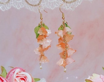 Puede incluir: Pendientes colgantes con racimos de flores color melocotón y detalles dorados. Cada pendiente tiene un gancho dorado y pequeñas hojas verdes. Los pendientes se presentan sobre un fondo rosa suave con detalles florales.