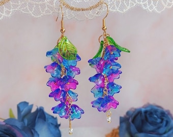 Puede incluir: Pendientes colgantes con racimos de cuentas en forma de flor azul y morada translúcida. Los pendientes tienen ganchos dorados, adornos de hojas verdes y pequeñas cuentas blancas. Fondo con rosas azules y encaje. Miden unos 8 cm.