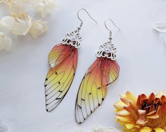 Puede incluir: Pendientes colgantes con diseño de ala de mariposa. Las alas van del amarillo al naranja y al rojo, con detalles de venas negras. Los pendientes tienen adornos de filigrana plateada con pequeños cristales transparentes.