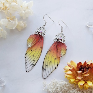 May include: A pair of dangle earrings featuring a butterfly wing design. The wings transition from yellow to orange and red, with delicate black vein detailing. The earrings have silver-colored filigree tops with small, clear crystals.