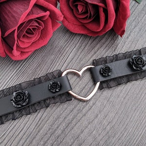 Könnte beinhalten: Schwarze Halskette mit silbernem Herz und schwarzen Rosen. Die Halskette hat einen schwarzen Spitzensaum und liegt auf einer grauen Holzoberfläche, mit zwei dunkelroten Rosen im Hintergrund.