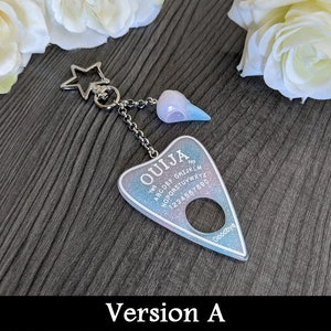 May include: A keychain featuring a Ouija board design. The planchette-shaped charm has a pastel blue and purple glitter background with the word "OUJIA" and other text. It is connected to a star-shaped charm and a small skull charm.