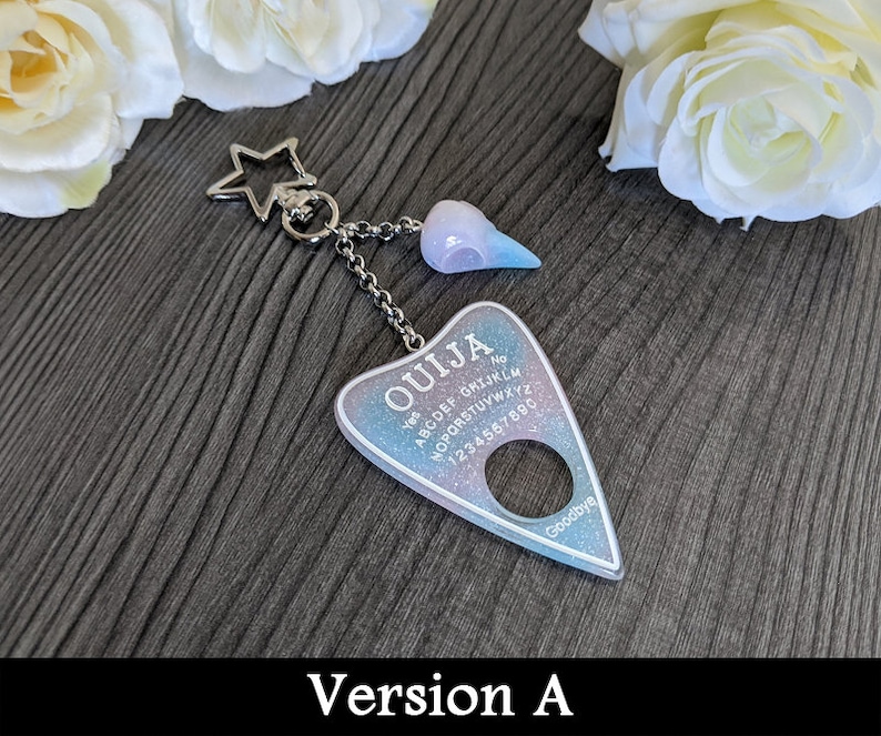May include: A keychain featuring a Ouija board design. The planchette-shaped charm has a pastel blue and purple glitter background with the word "OUJIA" and other text. It is connected to a star-shaped charm and a small skull charm.