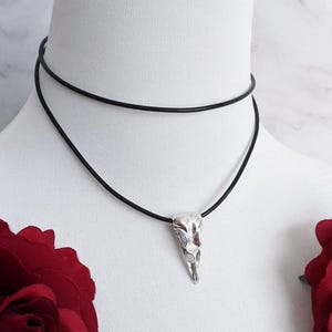 Könnte beinhalten: Halskette mit silbernem Anhänger in Form eines Rabenschädels. Der Anhänger hängt an einem schwarzen Kordel-Choker. Die Halskette wird auf einer weißen Schaufensterpuppe mit roten Rosen im Vordergrund präsentiert.