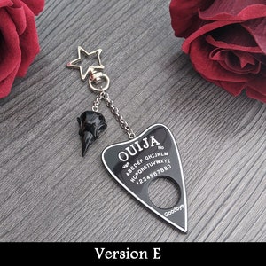 May include: A black Ouija board keychain with a raven skull charm. The planchette-shaped board has white lettering and numbers, with "Ouija" at the top. A silver star-shaped clasp and chain connect the charms.