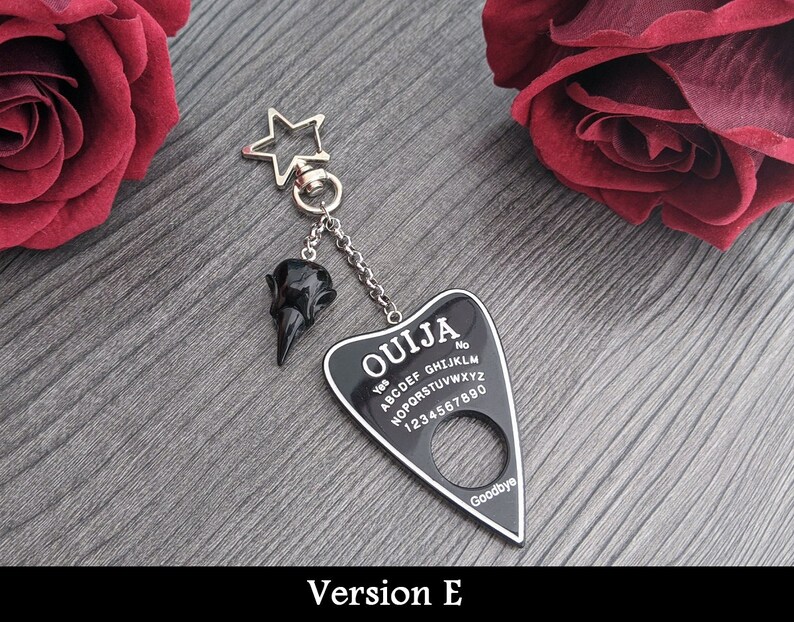 May include: A black Ouija board keychain with a raven skull charm. The planchette-shaped board has white lettering and numbers, with "Ouija" at the top. A silver star-shaped clasp and chain connect the charms.