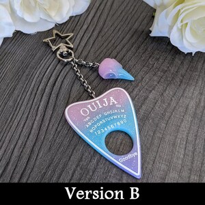 May include: A keychain featuring a Ouija board design in shades of purple, blue, and white with the word "OUJIA" at the top. It includes a star-shaped clasp and a charm shaped like a skull. The text "Version B" is at the bottom.