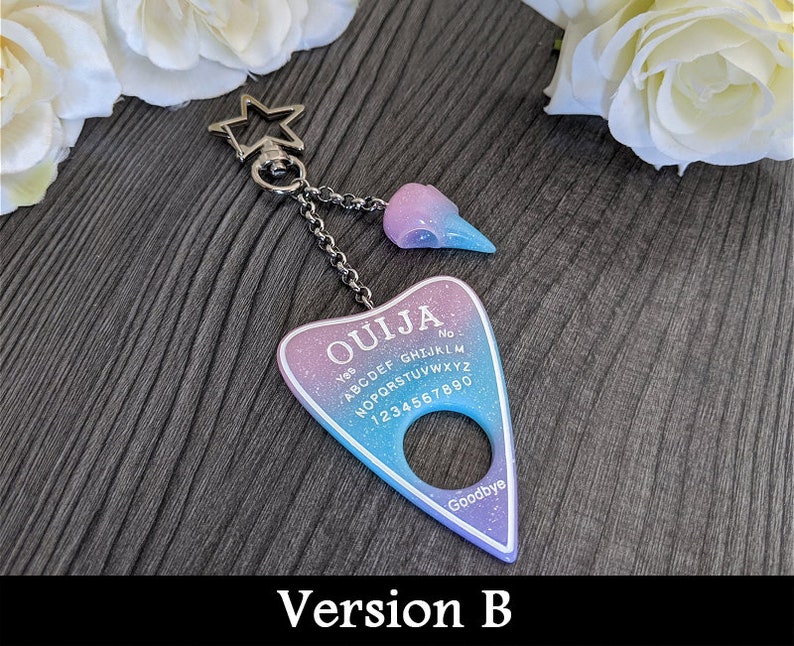 May include: A keychain featuring a Ouija board design in shades of purple, blue, and white with the word "OUJIA" at the top. It includes a star-shaped clasp and a charm shaped like a skull. The text "Version B" is at the bottom.