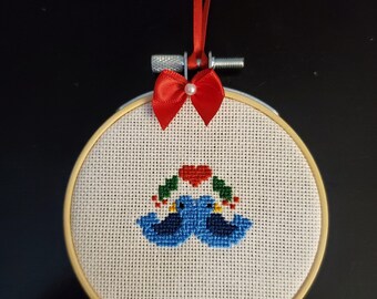 May include: A white embroidery hoop with a cross-stitch design of two blue birds with a red heart and green holly. The hoop is attached to a red ribbon.