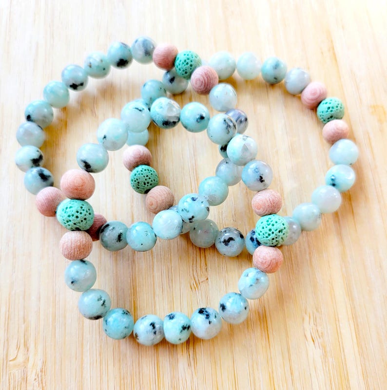 May include: Three beaded bracelets with light blue and speckled beads, interspersed with pink and green beads. The bracelets are arranged on a light wood surface, showcasing the jewelry's colors and textures.