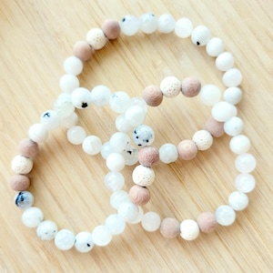 May include: Three beaded bracelets. The bracelets feature white, speckled, and light brown beads. The beads are strung together to form a circular shape. The bracelets are arranged on a light brown wooden surface.