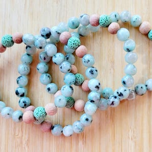May include: Four beaded bracelets with a mix of light blue, speckled, and wooden beads. The bracelets are arranged on a light brown wooden surface. The beads are round and vary in size, creating a textured look.