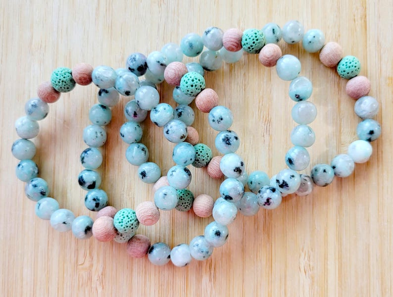 May include: Four beaded bracelets with a mix of light blue, speckled, and wooden beads. The bracelets are arranged on a light brown wooden surface. The beads are round and vary in size, creating a textured look.