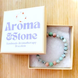 May include: An open box displaying a beaded bracelet. The bracelet features a mix of light green, pink, and white beads. The box is cream-colored with the words "Aroma & Stone" and "Handmade Aromatherapy Bracelets" printed on the lid.