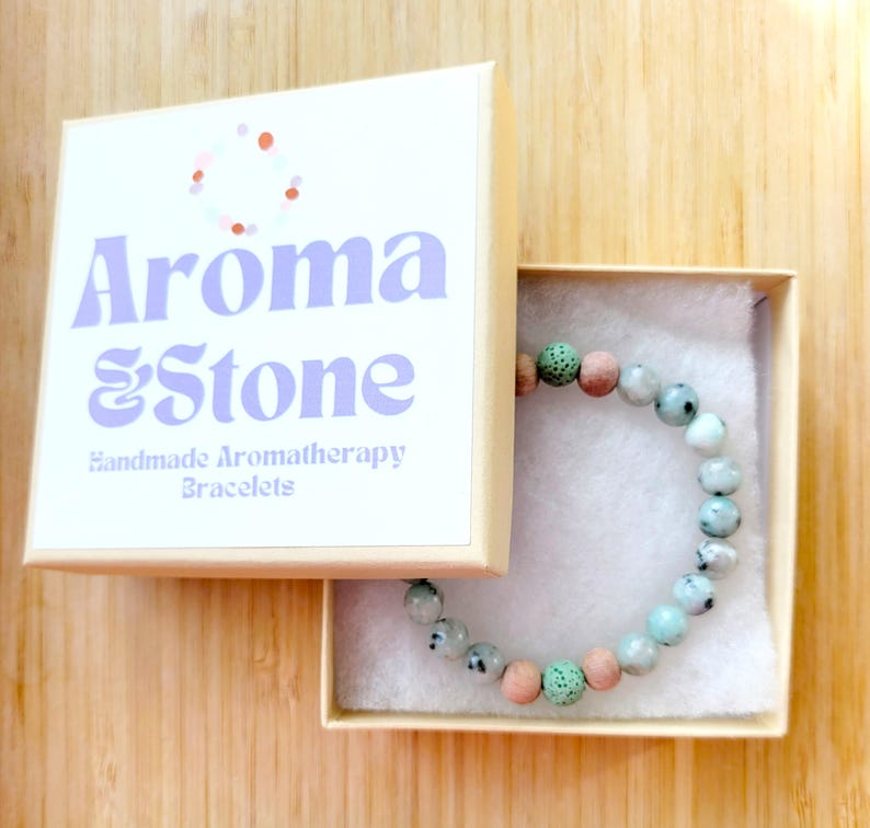 May include: An open box displaying a beaded bracelet. The bracelet features a mix of light green, pink, and white beads. The box is cream-colored with the words "Aroma & Stone" and "Handmade Aromatherapy Bracelets" printed on the lid.