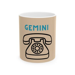 May include: A beige ceramic mug featuring the word GEMINI printed in blue capital letters above a black line drawing of a vintage rotary telephone with a handset resting on the base and a circular dial pad in the center of the device.
