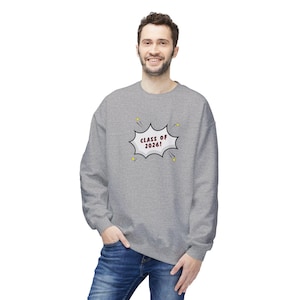 May include: A gray sweatshirt with the text "CLASS OF 2026!" in a speech bubble design. The sweatshirt is a crew neck style and is being worn by a person. The person is wearing blue jeans.