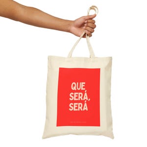 May include: A white canvas tote bag with red text that says "QUE SERÁ, SERÁ" in a bold, sans-serif font.