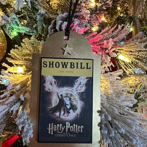 May include: Gold-colored Christmas ornament shaped like a playbill for the Harry Potter and the Cursed Child play. The ornament features a black and white image of the play's characters and the title "Harry Potter and the Cursed Child".