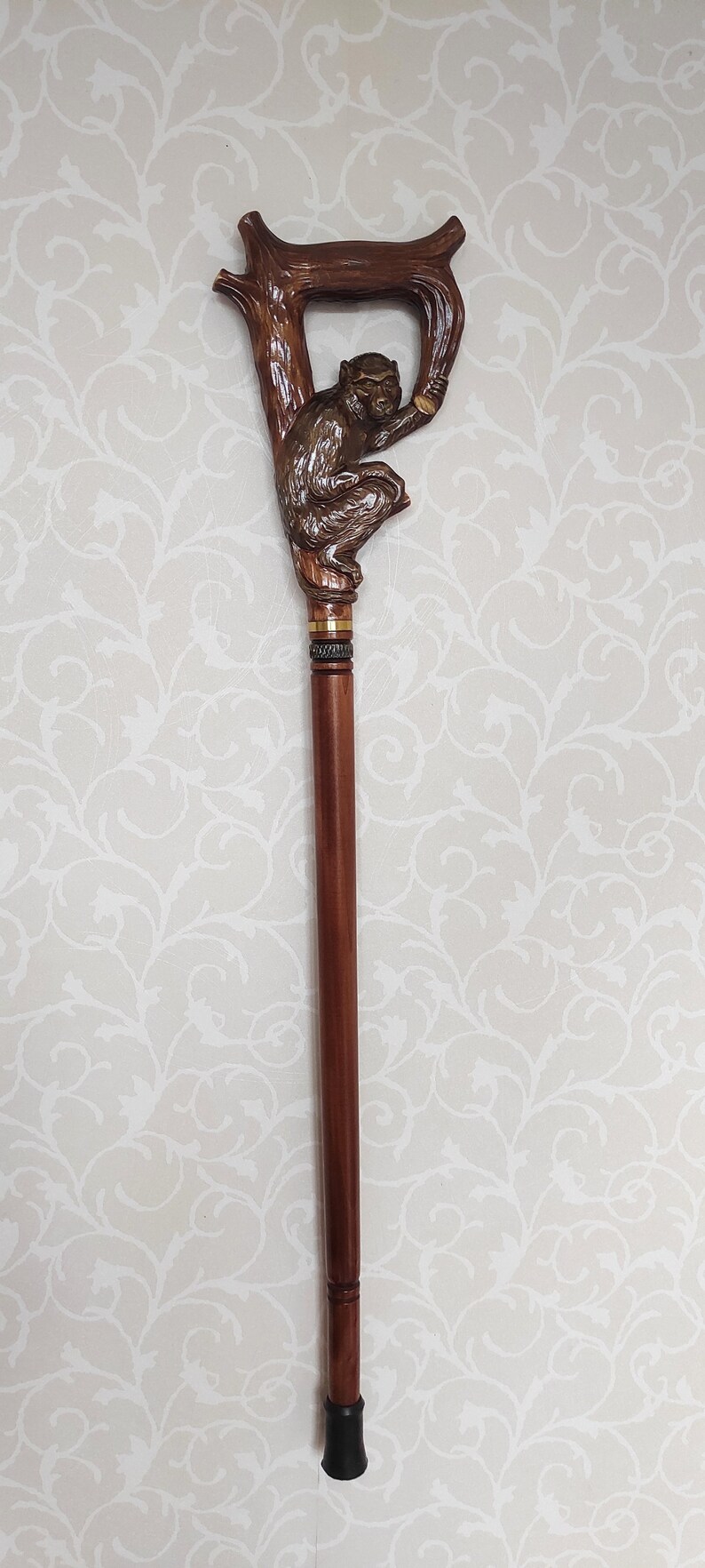 May include: A wooden walking stick with a carved monkey figure at the top. The monkey is brown and is holding onto a branch. The stick is brown and has a black rubber tip.