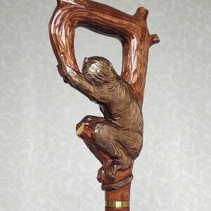 May include: A wooden walking stick with a carved monkey holding onto a branch. The stick has a brown stain and a gold band around the top.