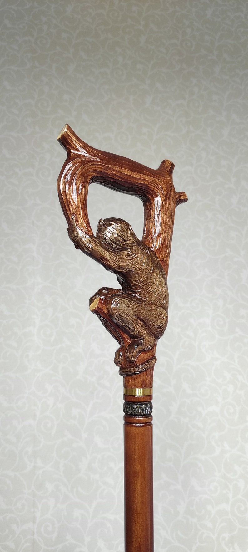 May include: A wooden walking stick with a carved monkey holding onto a branch. The stick has a brown stain and a gold band around the top.