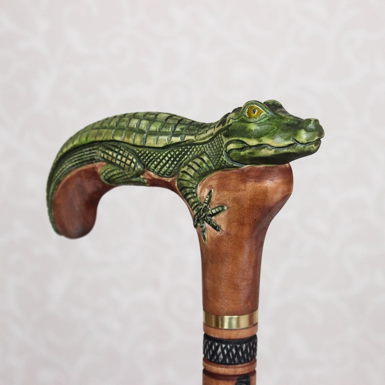 May include: A wooden walking stick with a carved alligator head handle. The alligator is green with a yellow eye and is positioned on top of the brown wooden handle.