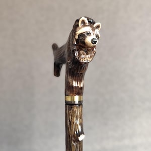 May include: A wooden walking stick with a carved raccoon head handle. The handle is brown and black with a gold band. The stick is brown and has a natural wood finish.
