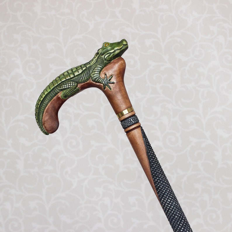 May include: A wooden walking stick with a carved alligator head handle. The handle is brown with green accents and the stick is black and brown.