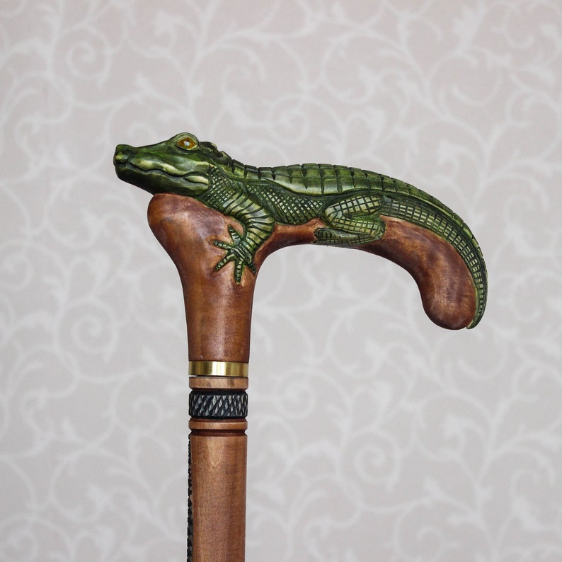 May include: A wooden walking stick with a carved alligator handle. The alligator is green and brown with a textured surface. The stick is brown and has a gold band and a black and white patterned band.