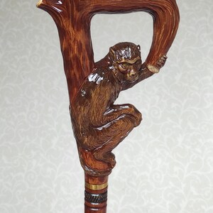 May include: A wooden walking stick with a carved monkey figure holding onto a branch. The stick has a brown finish and a gold band.