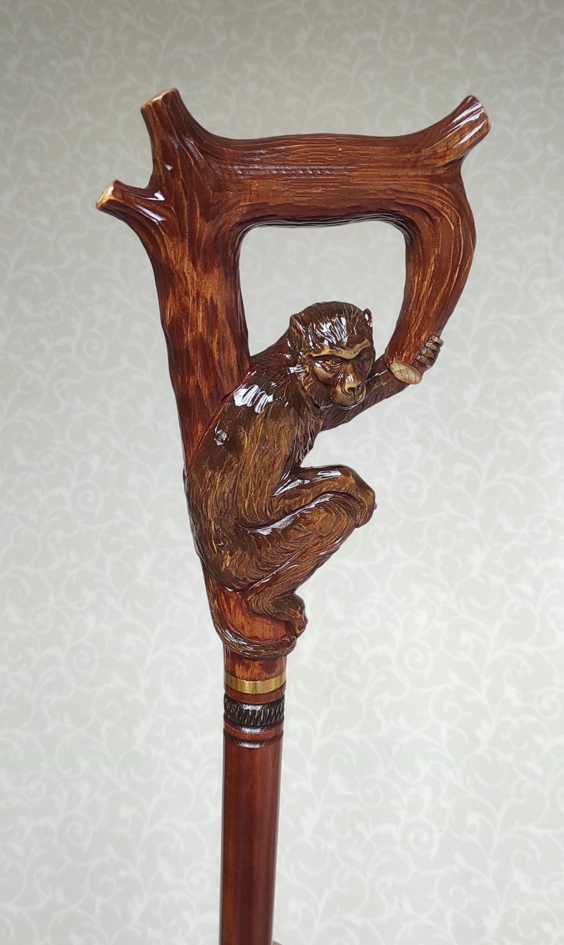 May include: A wooden walking stick with a carved monkey figure holding onto a branch. The stick has a brown finish and a gold band.